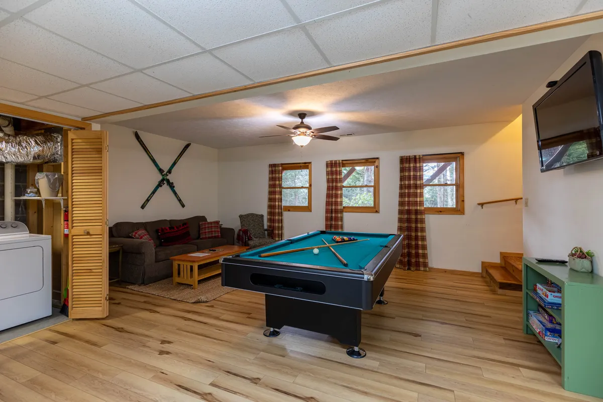 A relaxed game room scene with a pool table, sofa, windows letting in natural light, and a wooden staircase off to the side. The space feels open and welcoming, inviting groups to gather for games or simply unwind together.