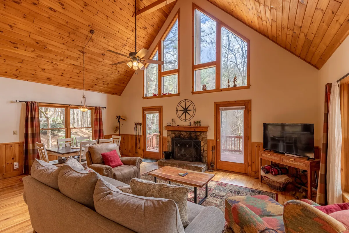 A spacious living area with a high ceiling and large windows that flood the room with sunlight. The focal point is a traditional wood-burning fireplace surrounded by comfortable seating, creating an inviting atmosphere for family gatherings or cozy nights by the fire. Natural elements and views of the outdoors are visible through the expansive windows.