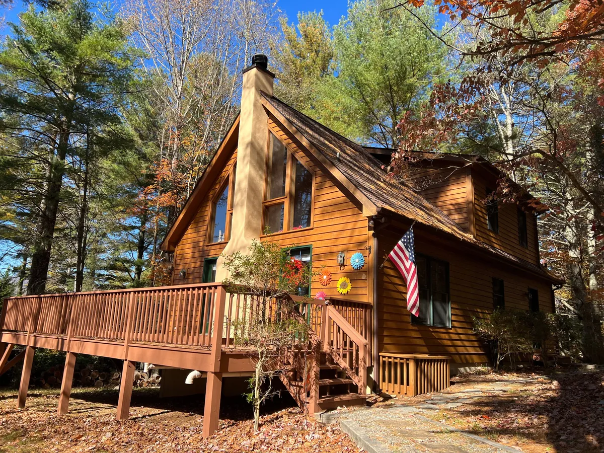 The image shows the exterior of a charming single-family cabin nestled in the Blue Ridge mountains, featuring a spacious wooden wrap-around deck adorned with colorful decorations and an American flag. Large windows invite natural light into the home, while the deck offers ample space for outdoor gatherings, relaxing, and enjoying stunning sunset views surrounded by nature.
