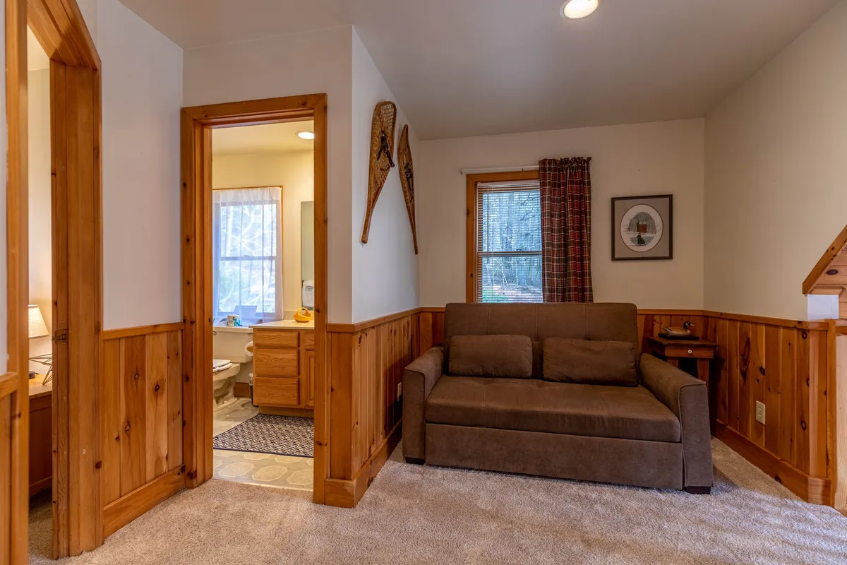 A welcoming loft area with a sofa, wood paneling, and a window with curtains, creating a cozy and versatile space for relaxation, lounging, or sleeping. Decorative touches and a bathroom door suggest functionality and comfort for groups.